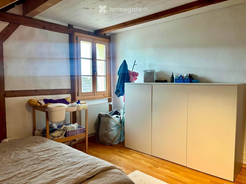 A cozy attic bedroom with wooden beams, a large window, a bed, a cabinet, a radiator, and various storage items including baskets and a small table.