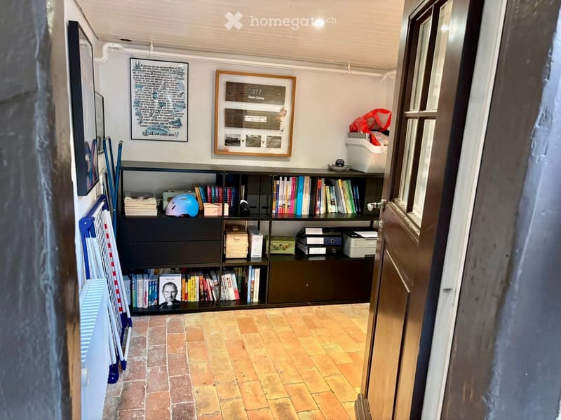 A small, well-organized room with a dark wooden bookshelf filled with books and various items, including a printer, a helmet, and storage boxes. The room features a tiled floor and a framed picture on the wall.