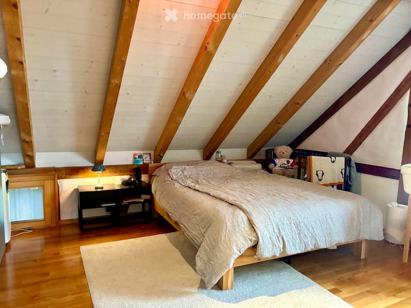 Cozy attic bedroom with wooden slanted ceilings, a large bed with beige bedding, a nightstand with a lamp, a mirror, and a small bookshelf with books and toys.