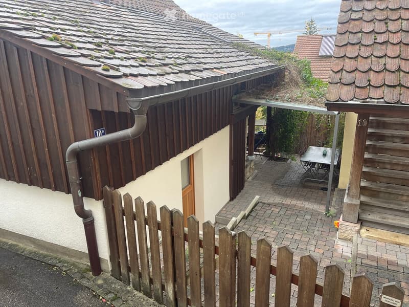 A small wooden house with a tiled roof, visible moss on the roof, and a small patio area with a table and chairs.