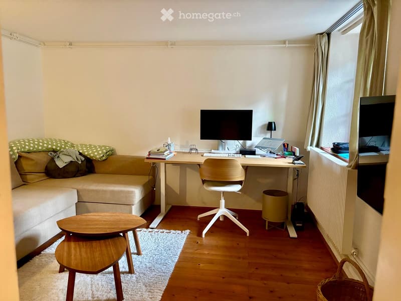 A small, cozy room with a sofa, a wooden table, a chair, a screen, and a lamp. The room has wooden flooring and a white rug.