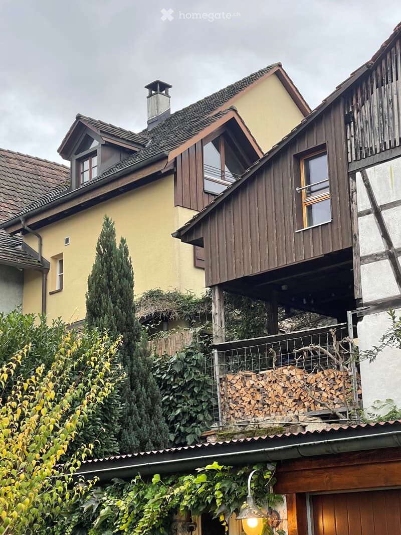 A multi-story house with a steeply pitched roof, wooden balcony, and chimney. The exterior features a mix of yellow and brown walls, with a wooden balcony and a stack of firewood on the roof.