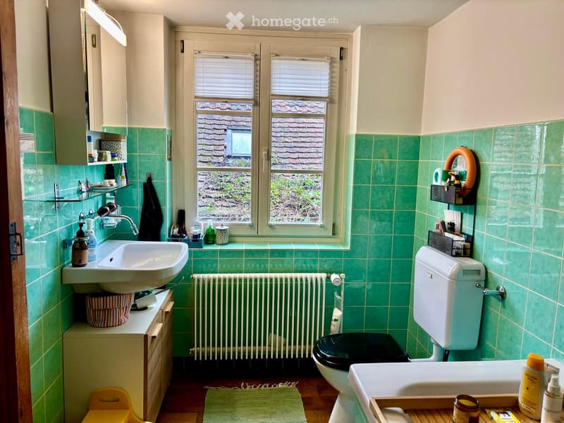 Bathroom with green tiled walls, a sink, toilet, bathtub, and a window overlooking a garden. Includes a radiator, shelves, and a small yellow stool.