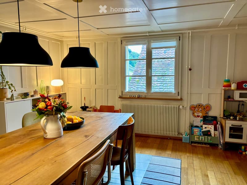 Bright kitchen with wooden table, chairs, vase with flowers, bowl of fruit, pendant lamps, and built-in cabinets.