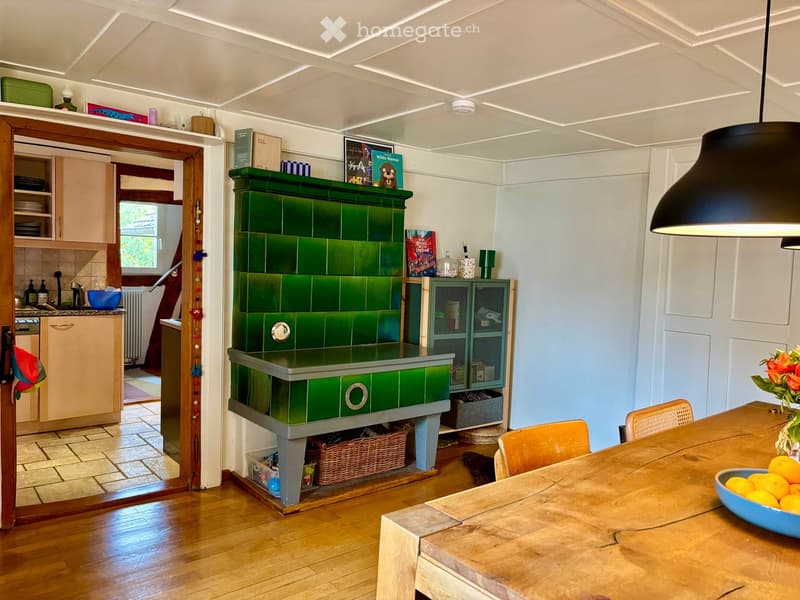 A cozy kitchen with a green tiled fireplace, wooden dining table, chairs, and various kitchen cabinets and appliances.