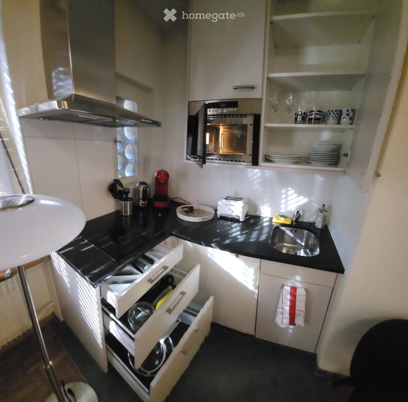 Modern kitchen with white cabinets, black countertops, stainless steel sink, and various appliances including a coffee maker, toaster, and oven. Open drawers reveal dishes and utensils.
