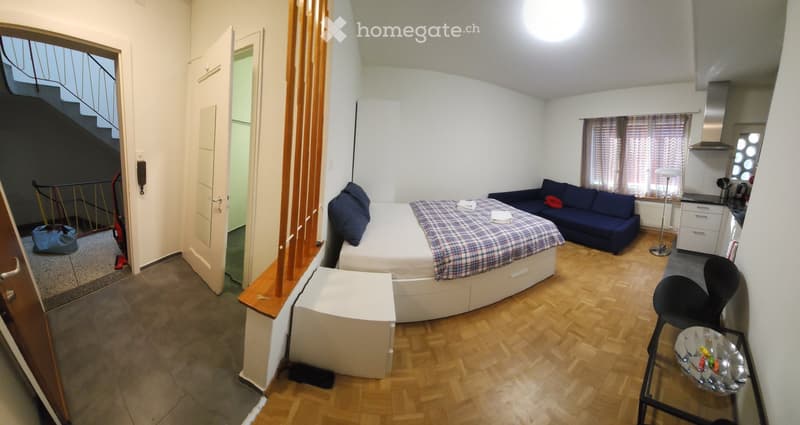 A compact, furnished room with a bed, sofa, chair, small table, and a kitchenette area with cabinets, sink, and appliances.