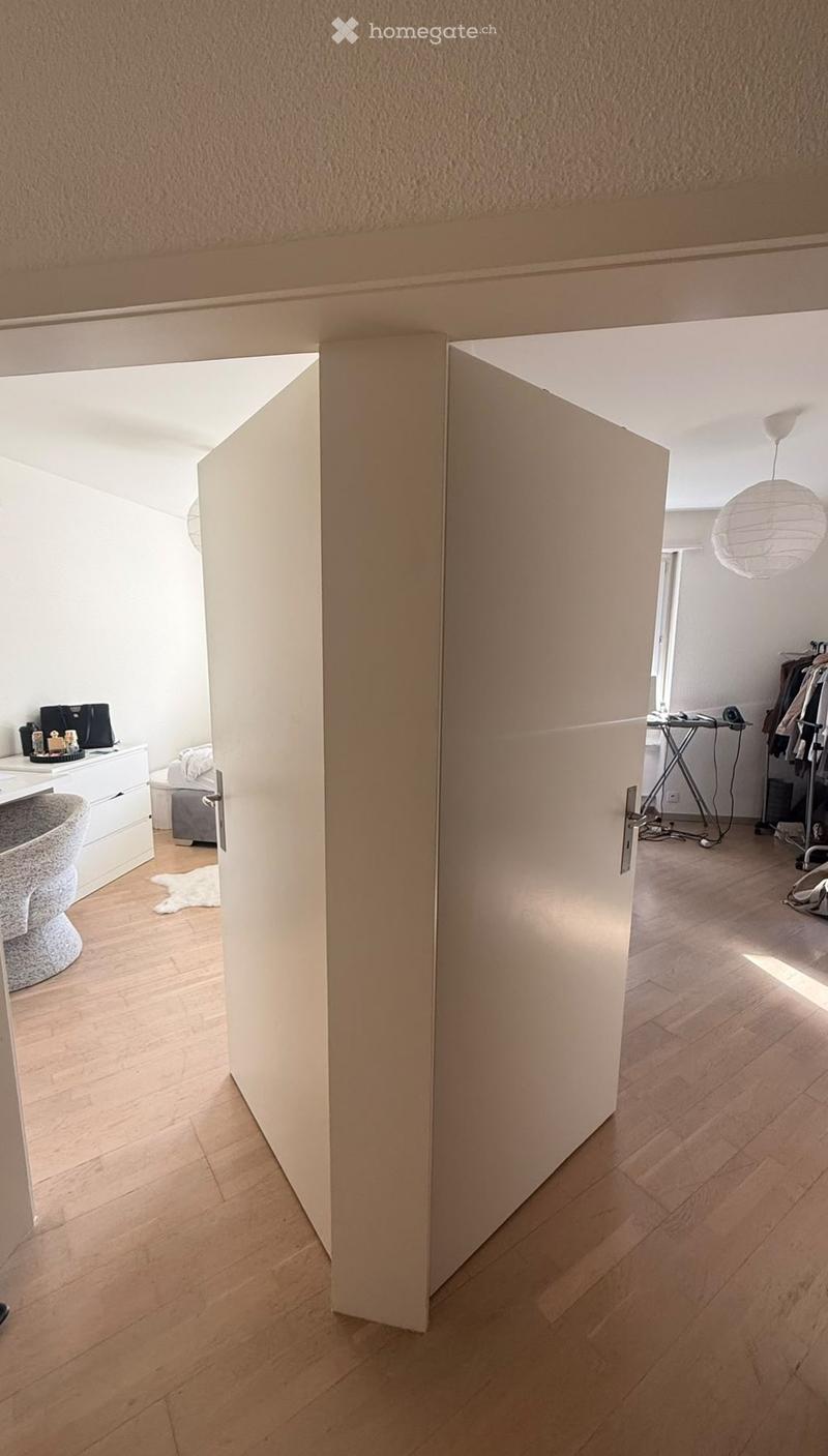 A modern hallway with light wooden flooring, white walls, and an open door leading to a room with a white desk, chair, and small rug.