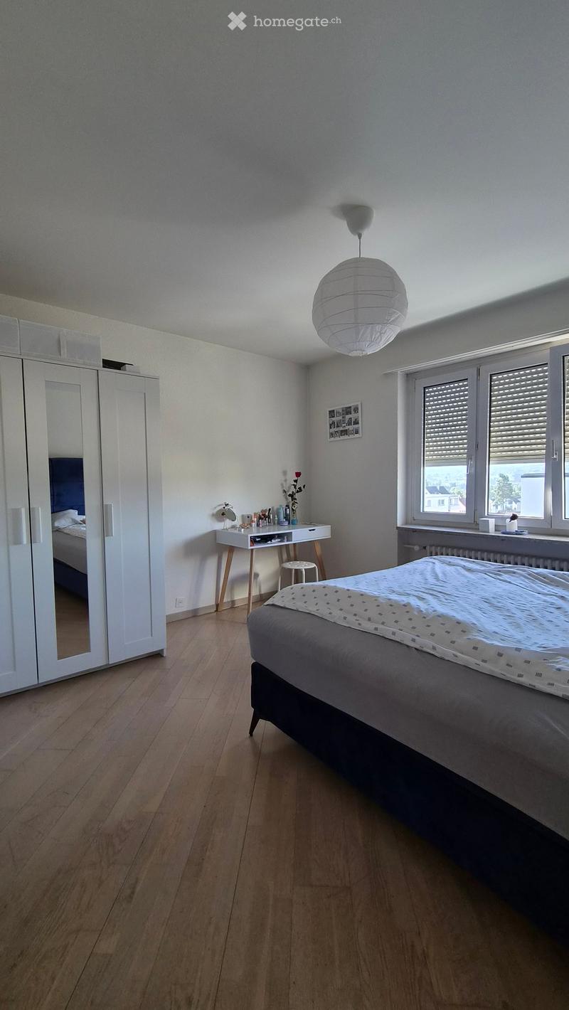 Modern bedroom with wooden flooring, a large bed with grey bedding, a white wardrobe with mirrored doors, a small desk with a chair, and a hanging paper lantern light fixture.