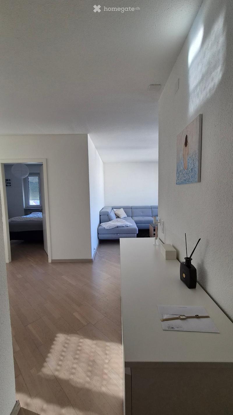 Bright hallway with wooden flooring, leading to a living area with a grey sofa and a bedroom with a bed visible in the background. A small table with decorative items is placed near the hallway entrance.