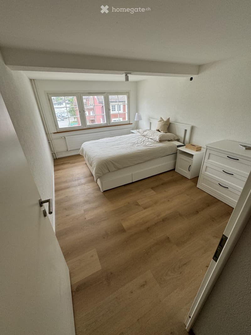 Bedroom with wooden flooring, a white bed with pillows, a nightstand with a lamp, and a white dresser with drawers.