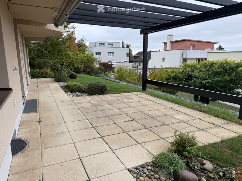 Balcony with tiled floor, covered by a wooden pergola, overlooking a landscaped garden with shrubs and small plants.