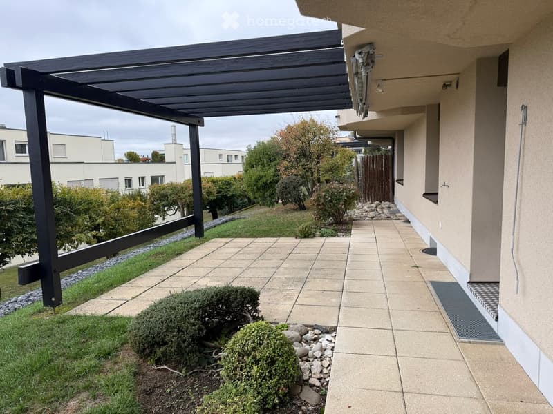 A covered patio area with a tiled floor, wooden pergola, and surrounding greenery, including bushes and trees.