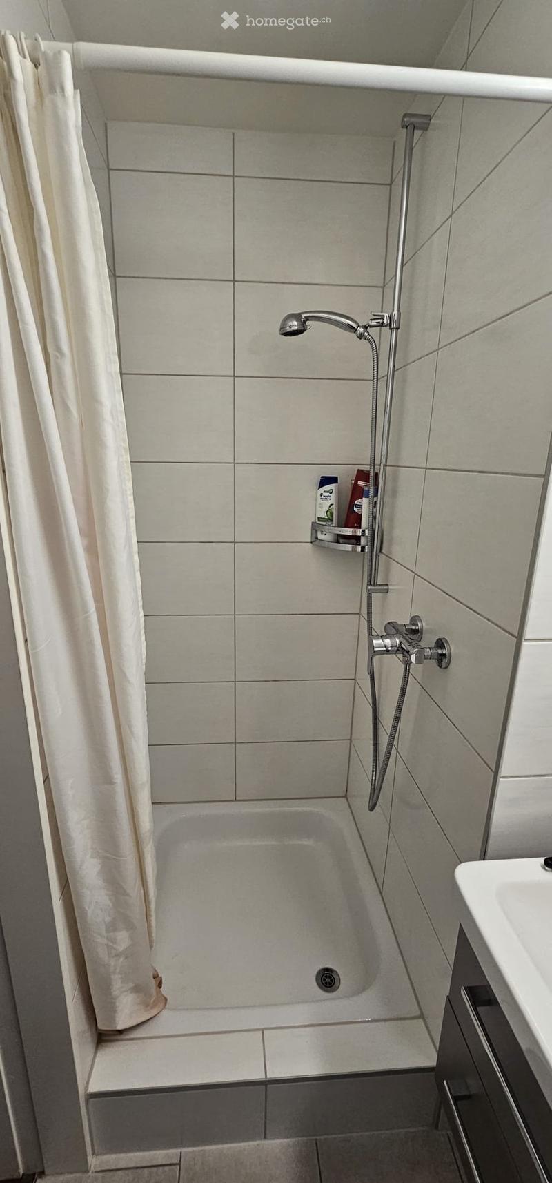 Modern bathroom with a bathtub, showerhead, and a small shelf holding toiletries. The shower curtain is beige, and the tiles are light grey.