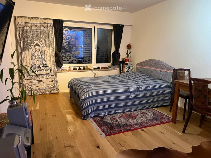 Cozy bedroom with wooden flooring, a bed with blue and white striped bedding, a small table with a chair, a curtain with a Buddha design, and a plant in a pot.