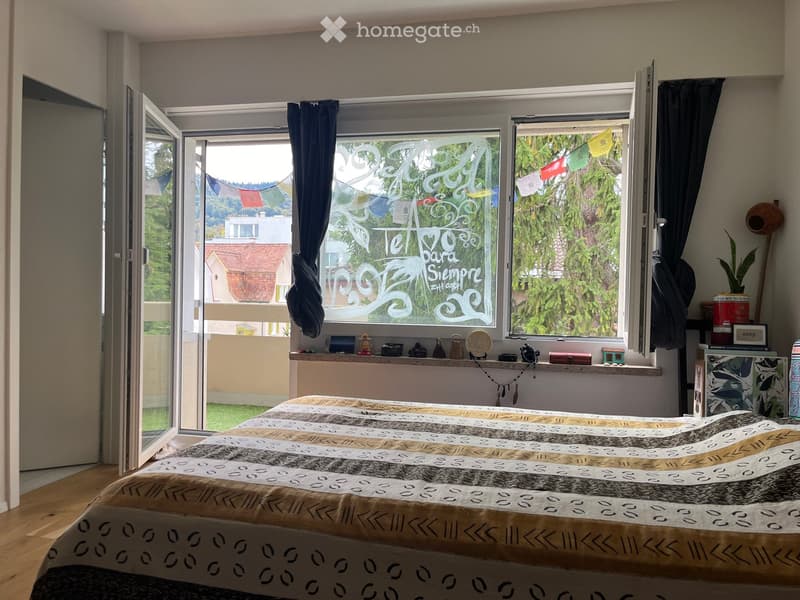 Cozy bedroom with a large bed, wooden floor, large windows with curtains, and a small table with decorative items.