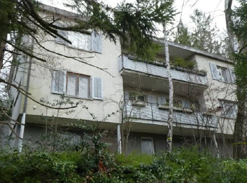 Multi-story residential building with balconies, surrounded by trees and greenery.