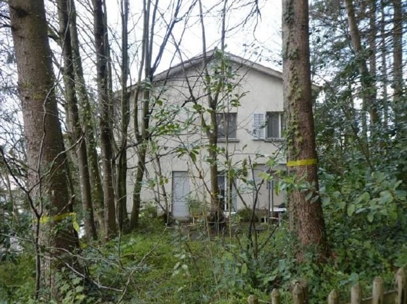 A two-story house with a beige exterior, surrounded by trees and greenery. The house features multiple windows and a visible front door.