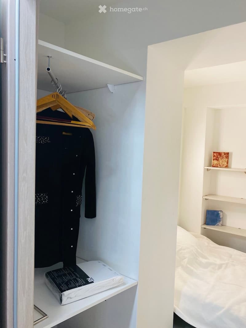 A compact bedroom with a neatly made bed, a built-in wardrobe with hanging clothes, and a small shelf holding folded items.