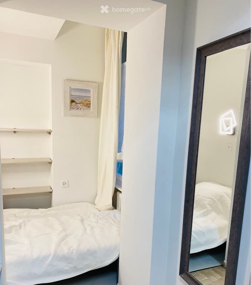 A compact bedroom with a neatly made bed, built-in shelves, a large mirror, and a framed picture on the wall.