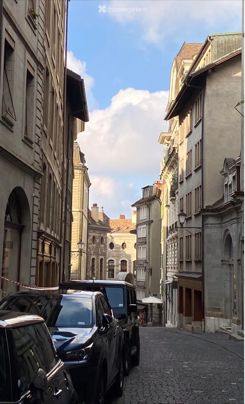 A narrow cobblestone street lined with multi-story buildings, featuring classic European architecture with ornate facades, balconies, and street lamps. A parked vehicle is visible on the left side.