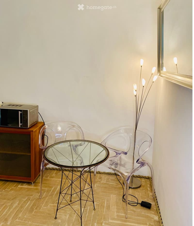 A small, modern room with a glass table, two transparent chairs, a microwave on a wooden stand, and a floor lamp with multiple bulbs.
