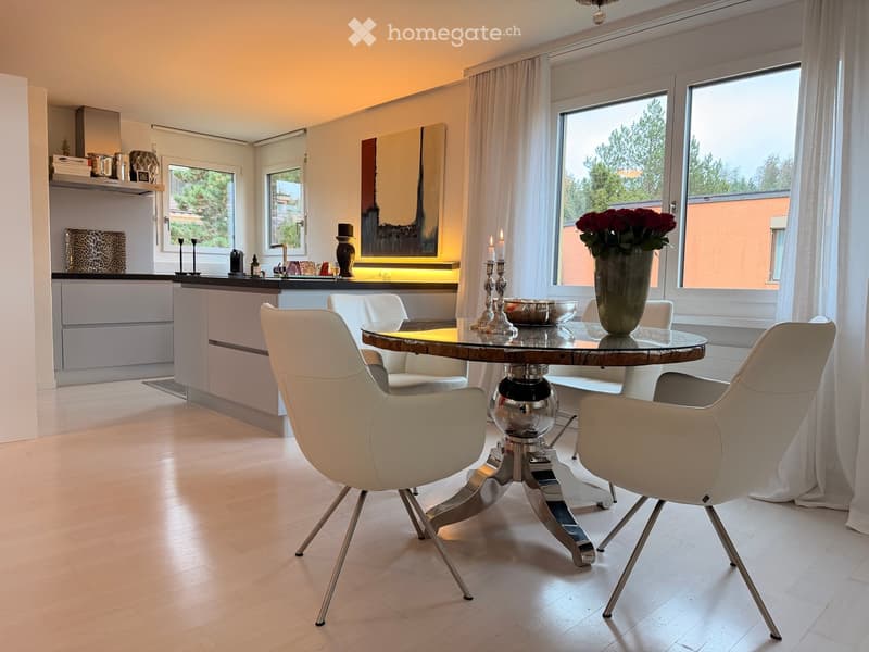 Modern dining area with a round glass table, white chairs, a vase with red flowers, and a sleek kitchen area in the background.