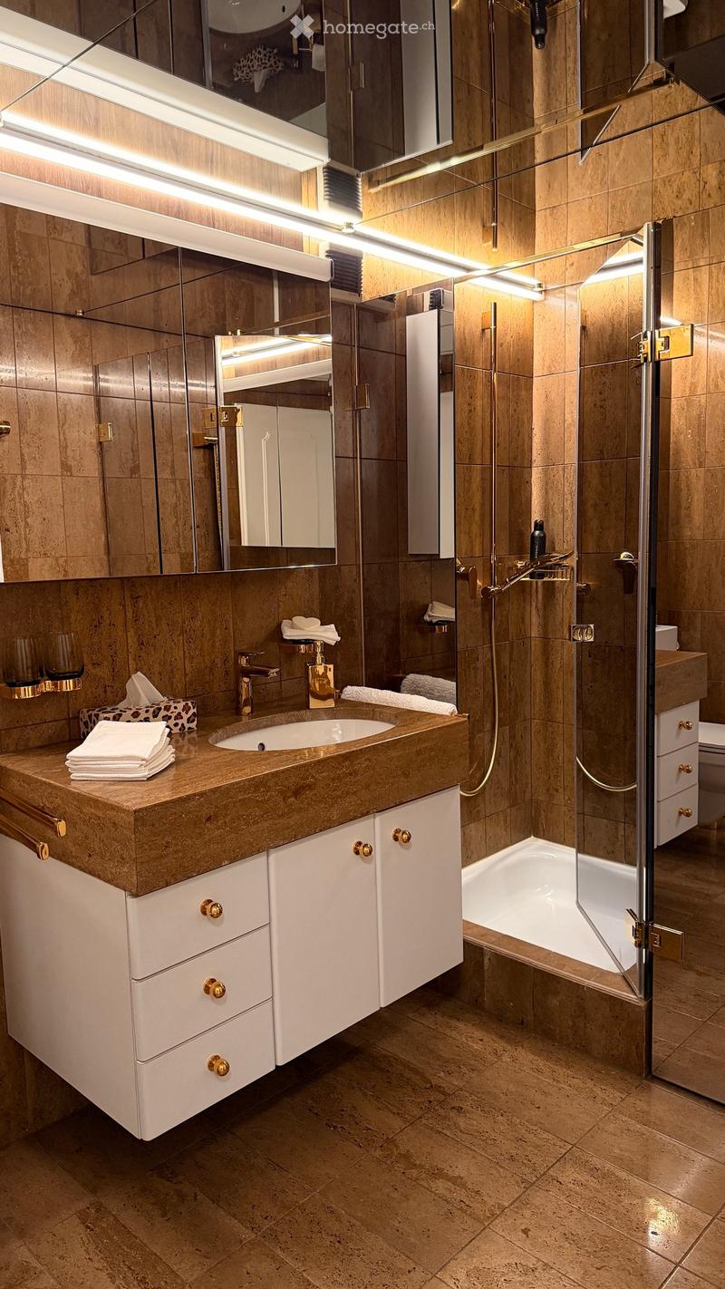 Modern bathroom with a large sink, mirrored cabinet, bathtub, and shower area. Features include a marble countertop, white cabinets, and gold fixtures.
