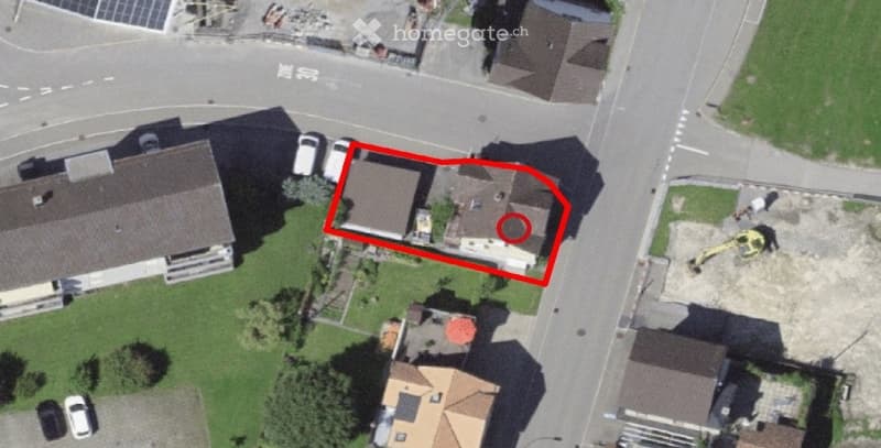 Aerial view of a residential building with a flat roof, surrounded by greenery and neighboring houses. The building appears to be a single-story structure with a small balcony or terrace area visible on the roof.