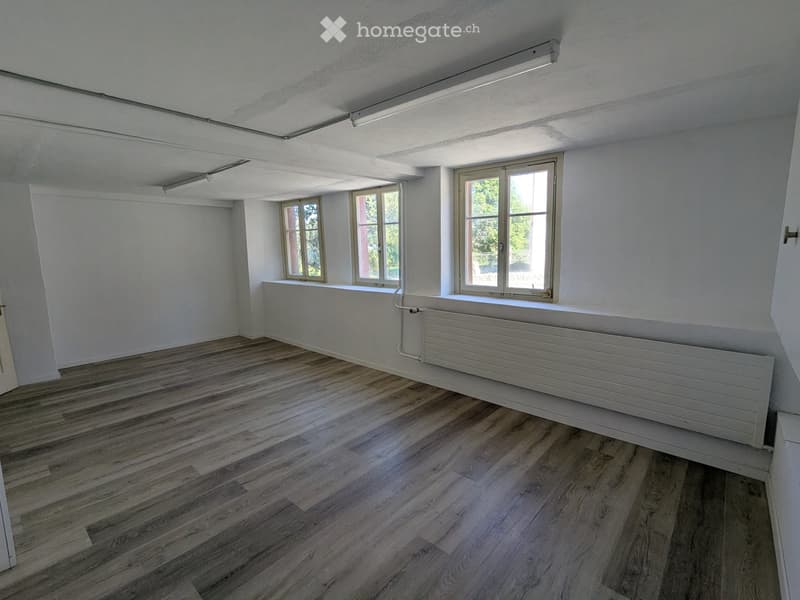 Empty room with wooden flooring, white walls, and large windows letting in natural light. Features a radiator and a door visible on the left side.