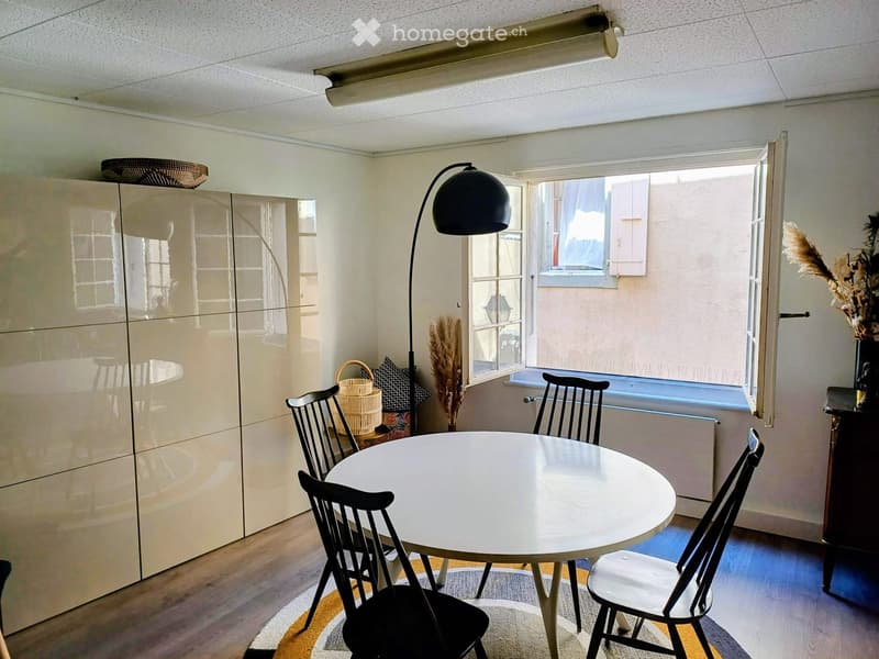 A modern, well-lit room with a round white table surrounded by black chairs, a floor lamp, a large cabinet, and decorative items including a wicker basket and a vase with dried flowers.