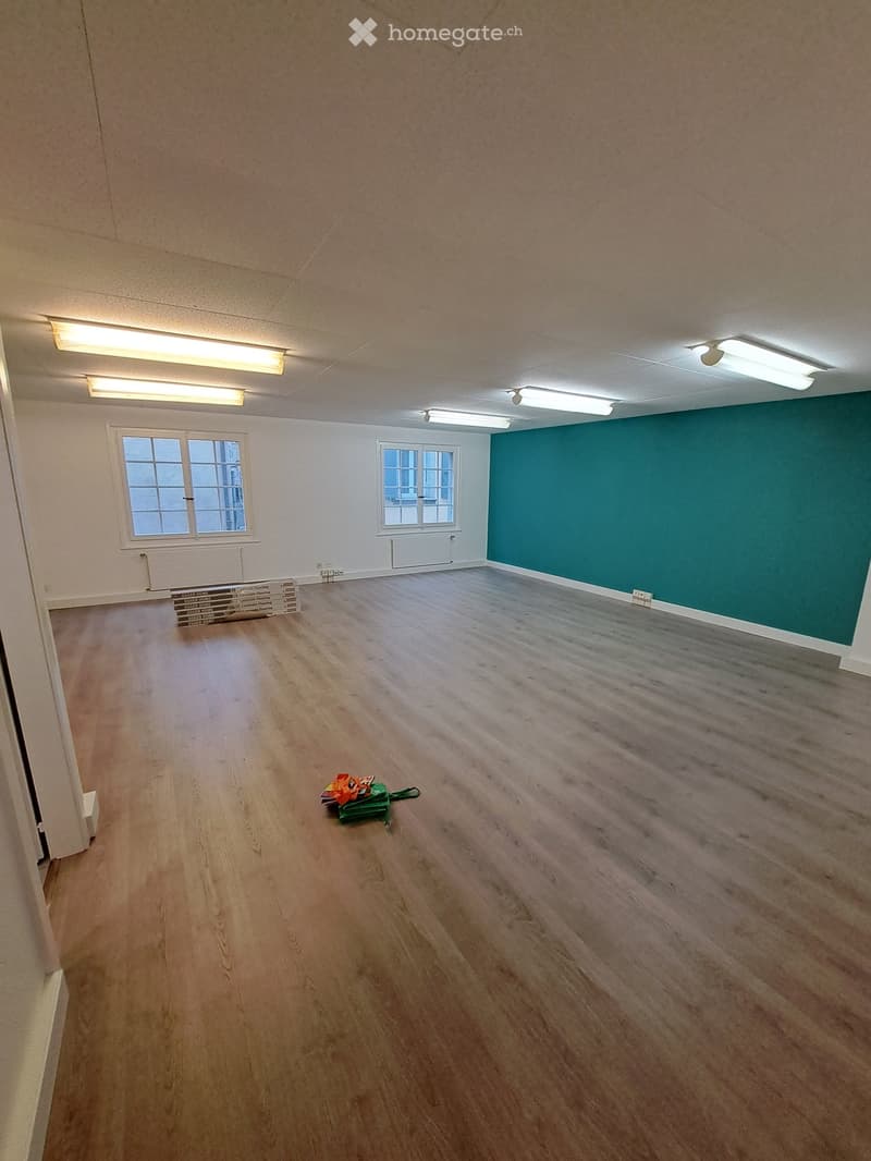 Empty room with wooden flooring, white walls, and two windows. There are some stacked boxes and a small green bag on the floor.
