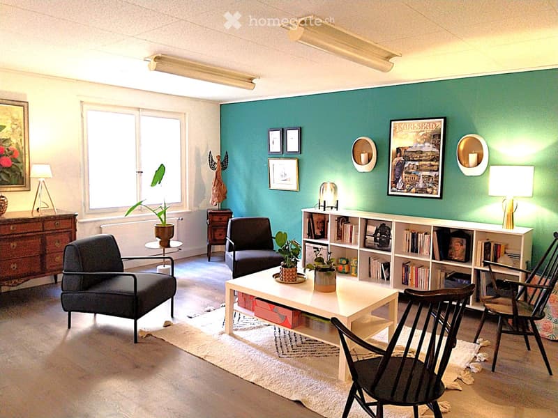 A modern living room with a teal accent wall, white table, two chairs, bookshelf, potted plants, and decorative items.