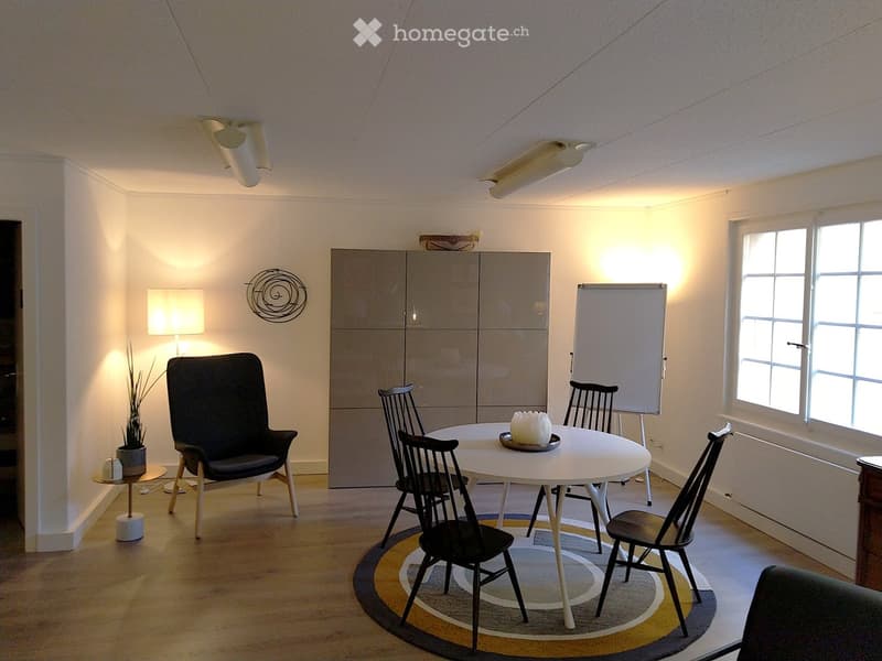 A modern, well-lit room with a round white table surrounded by black chairs, a black armchair, a floor lamp, a whiteboard, and a cabinet with a decorative wall piece above it.