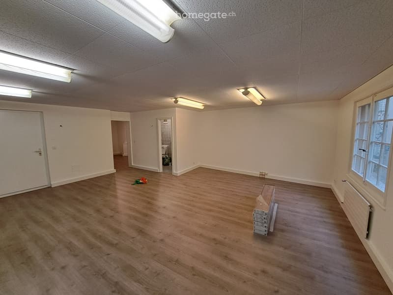 Empty room with wooden flooring, white walls, and fluorescent lighting. There are doors leading to other rooms, a window, and some stacked boxes in the corner.