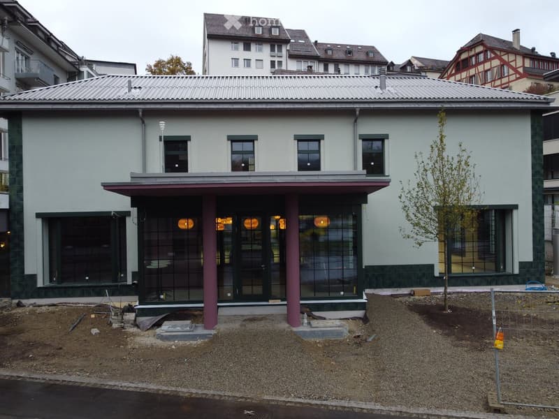 Modern two-story building with light gray facade, large windows, and a flat roof. The entrance features a covered porch with glass doors and pink columns. The surrounding area includes a small tree and construction materials.
