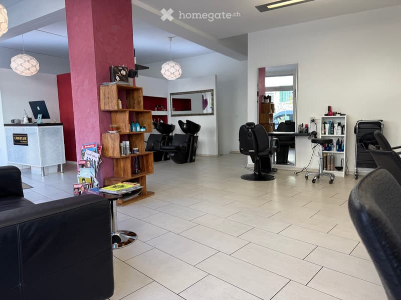 Modern, well-lit salon with white tiled floors, a reception desk, multiple salon chairs, shelves with hair products, and a comfortable seating area with a sofa and table.