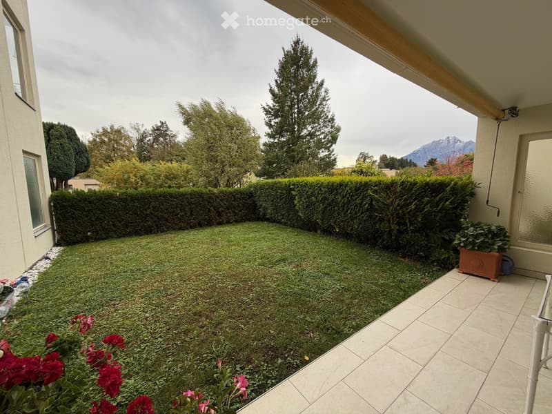 A well-maintained garden with a neatly trimmed hedge, a small lawn, and a variety of flowers and plants. The area is bordered by a tiled patio and features a scenic mountain view in the background.