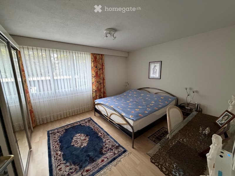 A furnished bedroom with a bed, a chair, a table, a picture frame, and a lamp. The room features wooden flooring, a large window with curtains, and a blue rug.