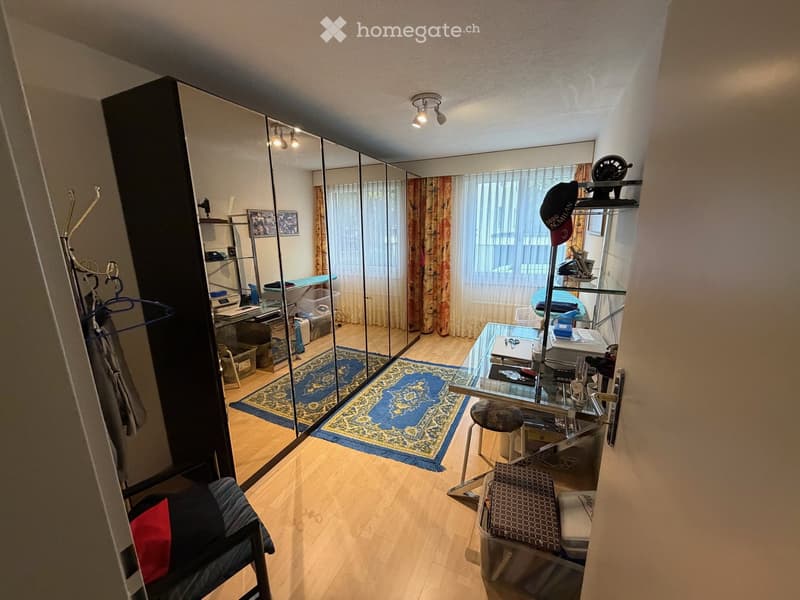 A furnished room with a large mirror, a chair, a small table, and a stool. The room has wooden flooring and a patterned rug.