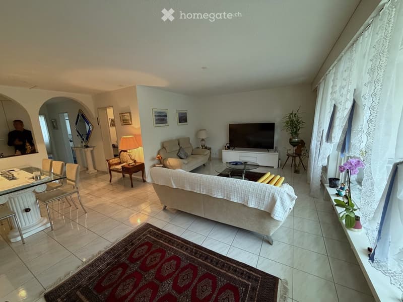 Bright living room with tiled flooring, large windows with white curtains, and a mix of seating including a sofa, armchair, and dining table with chairs. A television is mounted on a white stand, and decorative elements such as lamps, a mirror, and artwor