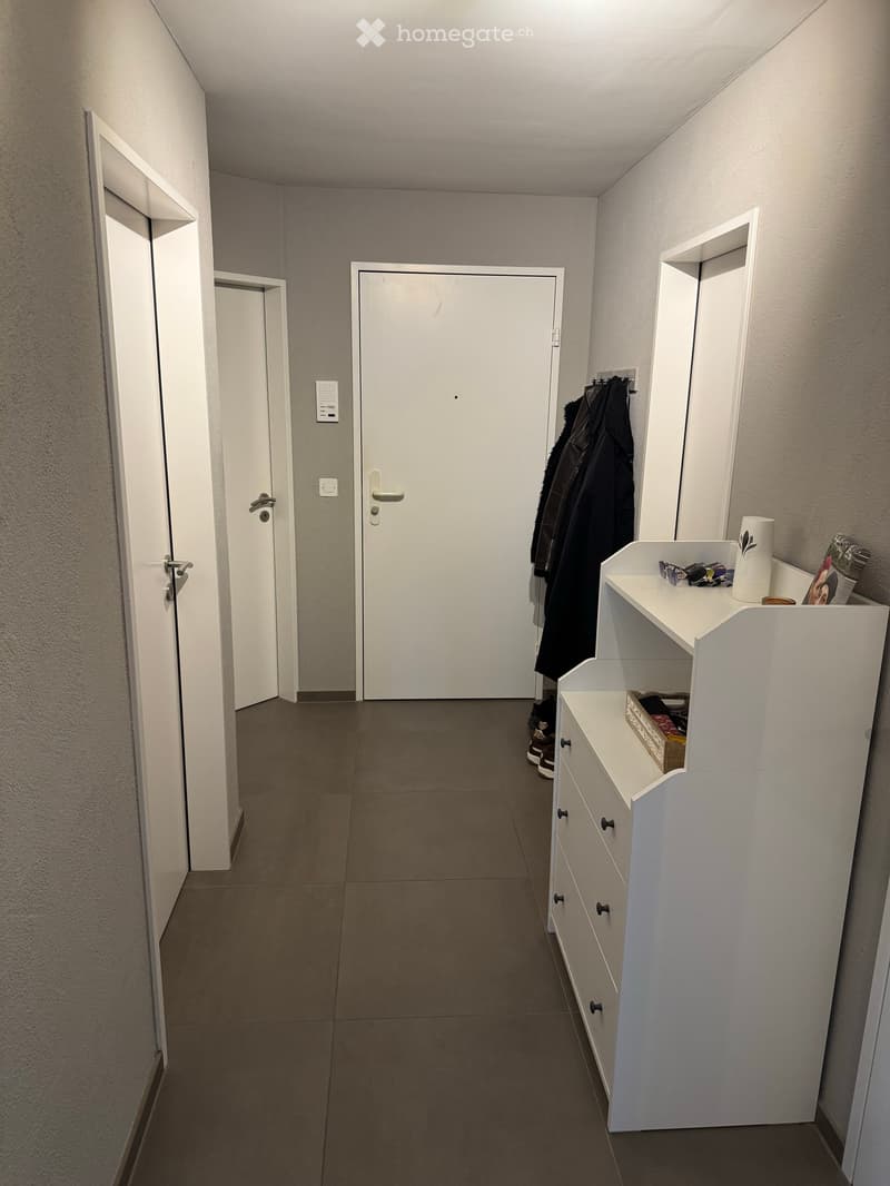 A clean, modern hallway with tiled flooring, white walls, and built-in storage units. A coat rack with jackets and shoes is visible, along with a small shelf holding various items.