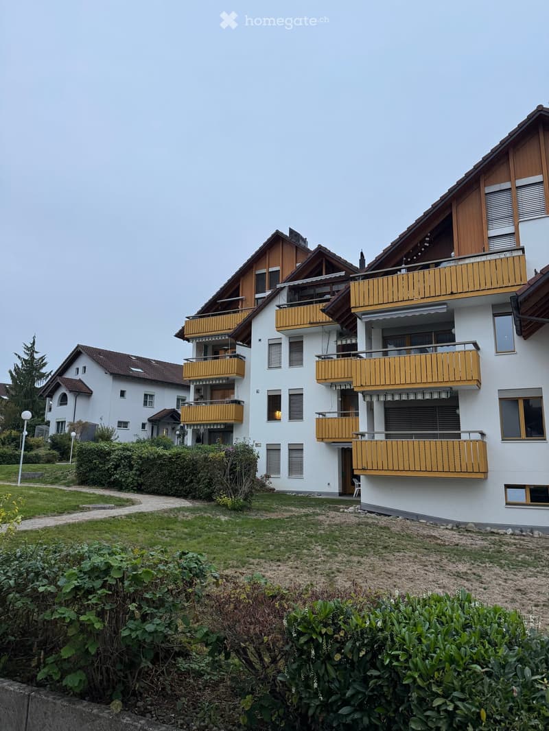 Multi-story residential building with white walls, wooden balconies, and a sloped roof. The building is surrounded by greenery and a paved pathway.