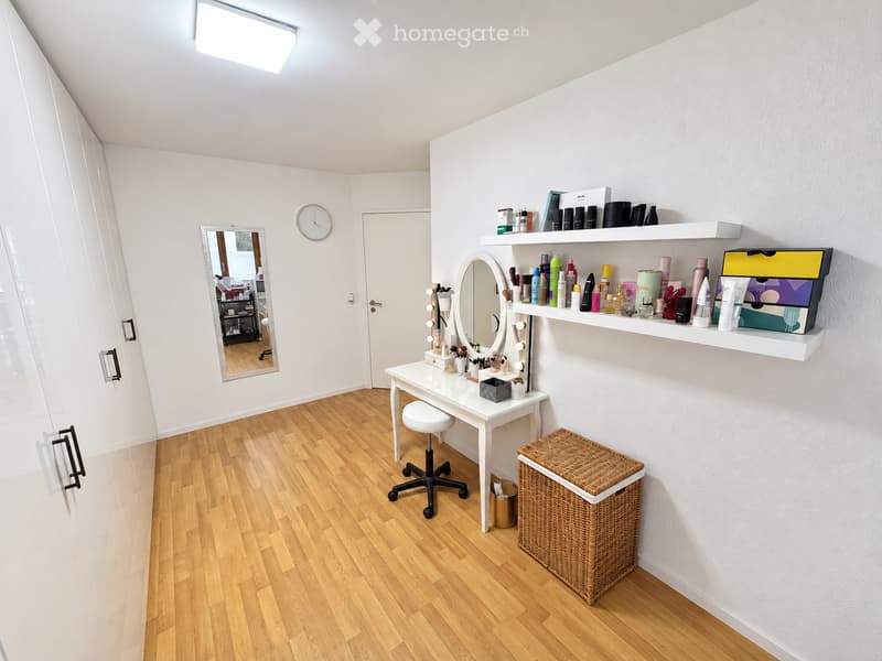 Well-lit vanity area with a white vanity table, round mirror, stool, shelves filled with cosmetics, and a wicker basket on the floor.