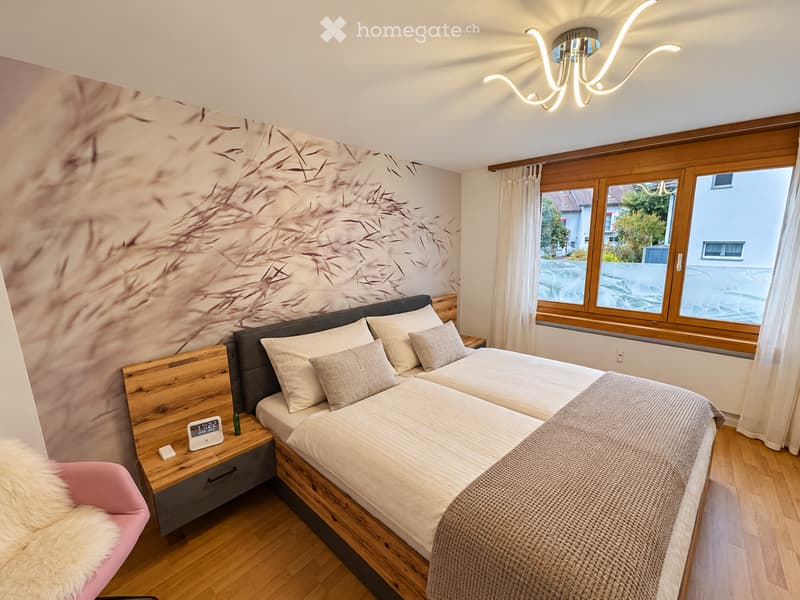 Cozy bedroom with wooden flooring, large window with white curtains, double bed with beige bedding, nightstand with clock, and a pink chair with a fur throw.