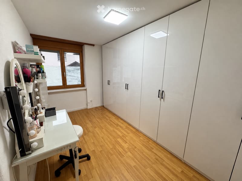 A well-lit room with wooden flooring, a large white cabinet, a vanity table with a mirror, and shelves holding various items.
