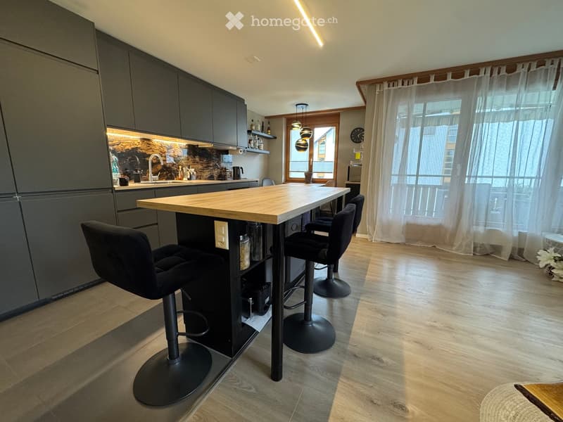 Modern kitchen with a central island, four black stools, sleek grey cabinets, and a large window with white curtains.