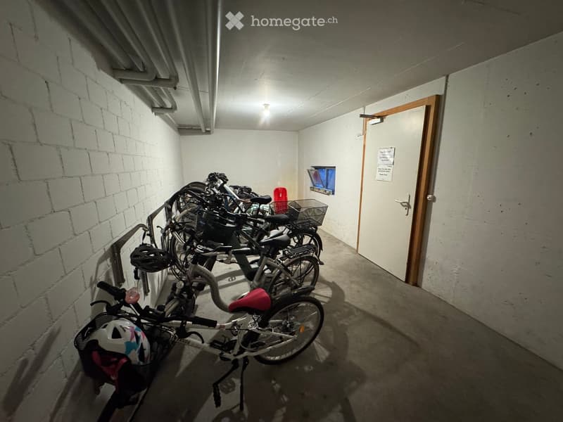 A small, concrete-walled room with several bicycles parked inside, helmets placed on the bikes, and a door leading out.