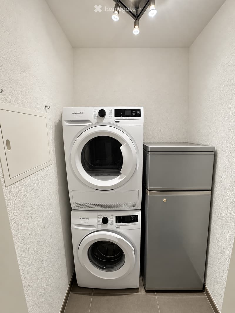 A small, well-lit room with a stacked washing machine and dryer unit, a refrigerator, and a cabinet.