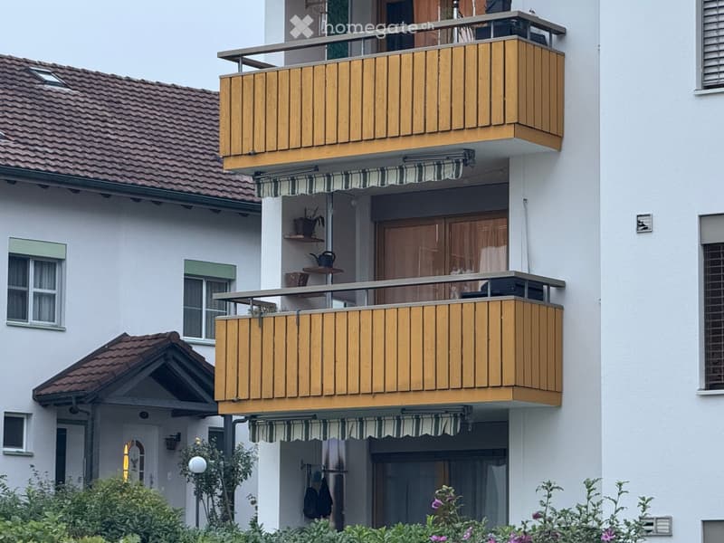Two-story residential building with multiple balconies, wooden railings, striped awnings, and small garden area.